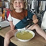 child, toddler, curly_hair, smiling, kitchen, countertop, bowl, whisk, batter, cooking, knife_rack, utensils, cabinet, appliance, clothing, happy, person, indoor, foot, table