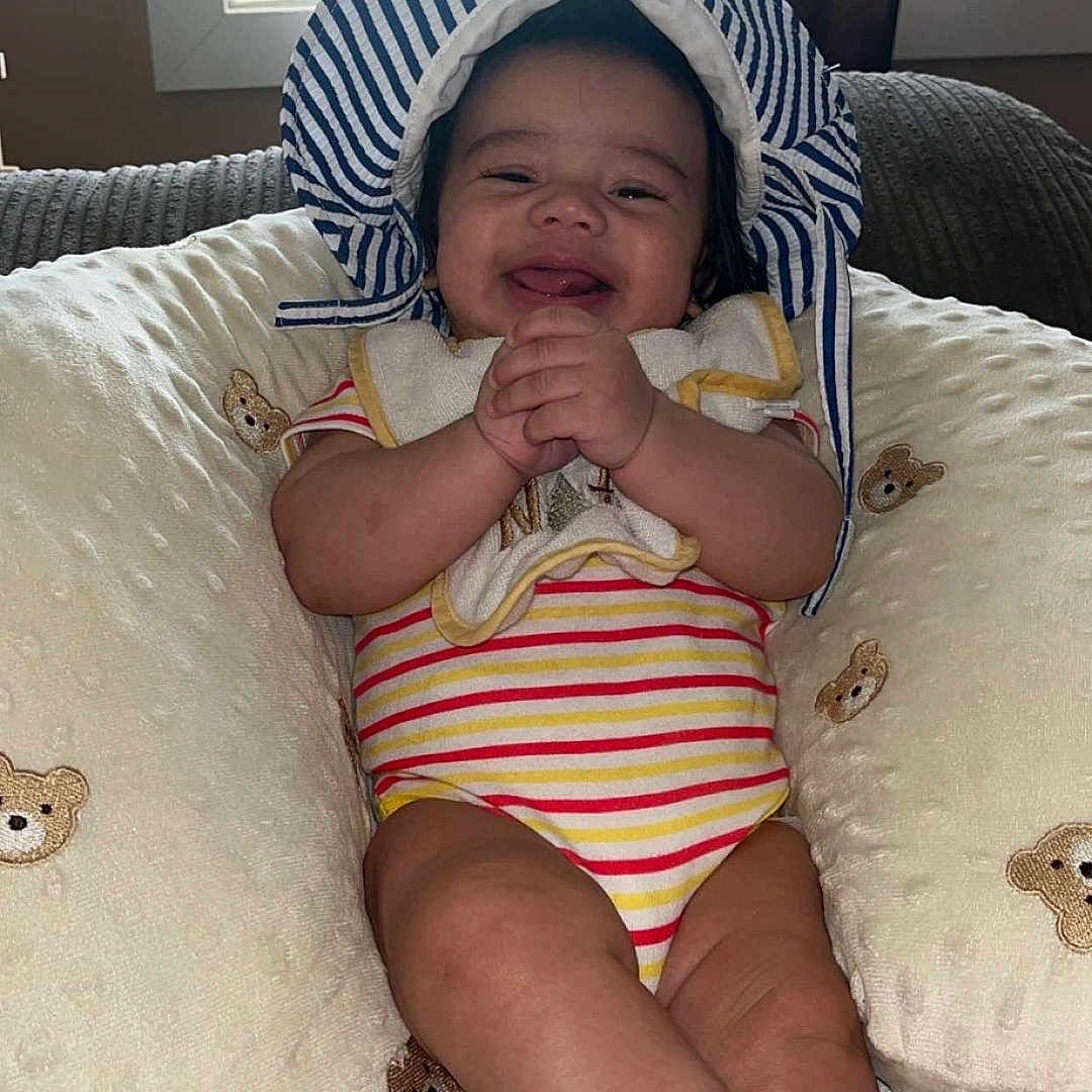 Rozay is registered to the contest to win money with this photo: baby, blanket, child, clapping_hands, cozy, curtain, cute, happy, hat, indoor, infant, person, pillow, portrait, red_socks, seated, smiling, striped_onesie, teddy_bear, window