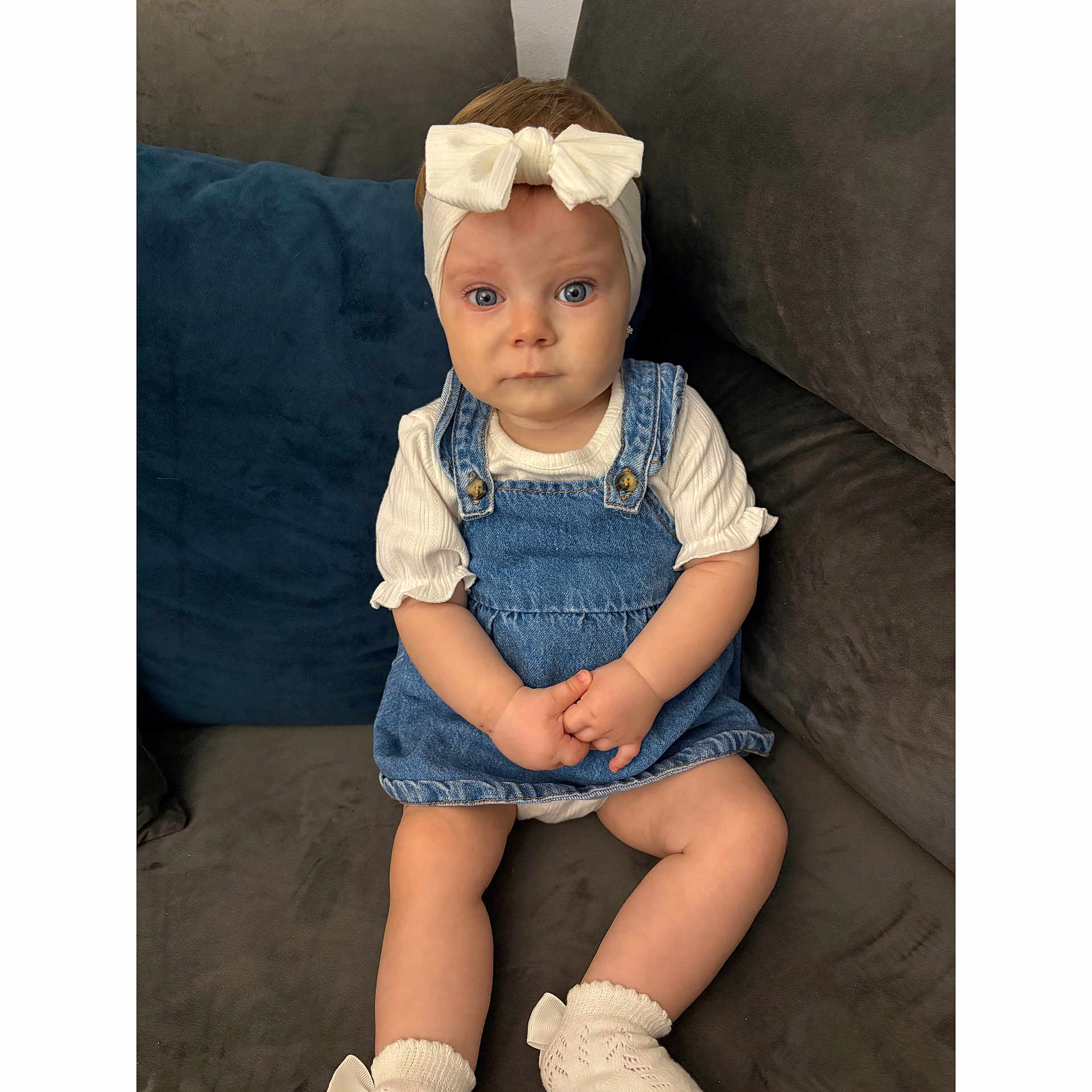 Ayla joined the competition — help win amazing prizes! baby, child, girl, denim_dress, headband, bow, socks, couch, blue_eyes, sitting, indoors, clothing, cute, portrait, hands, furniture, expression, soft_lighting, white, cozy
