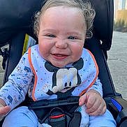 Mathias is registered to the contest to win money with this photo: baby, smiling, stroller, bib, mickey_mouse, curly_hair, pajamas, outdoor, happy, child, infant, seatbelt, blanket, hand, face, person, cute, daylight, expression, playful