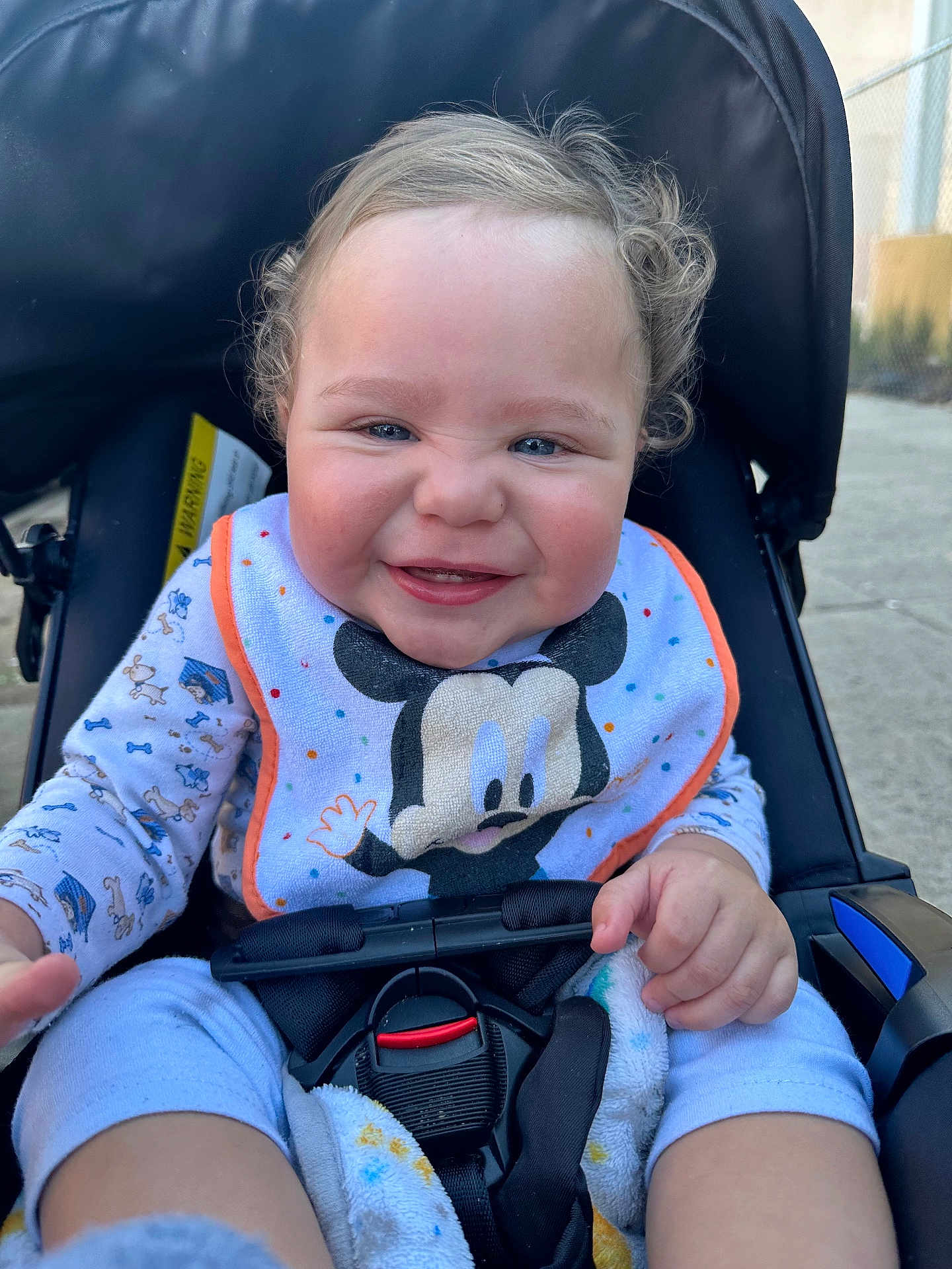 Mathias is registered to the contest to win money with this photo: baby, smiling, stroller, bib, mickey_mouse, curly_hair, pajamas, outdoor, happy, child, infant, seatbelt, blanket, hand, face, person, cute, daylight, expression, playful