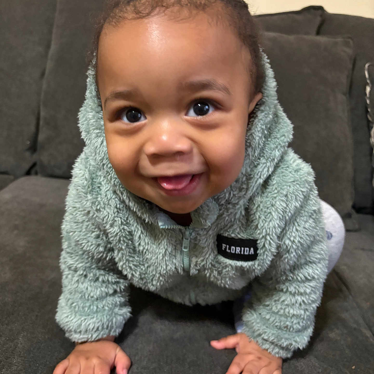 Azaire is registered to the contest to win money with this photo: baby, child, closeup, couch, cozy, crawling, curious, cute, face, furniture, fuzzy_jacket, green_clothing, hands, happy, home, indoor, person, portrait, smiling, young_child