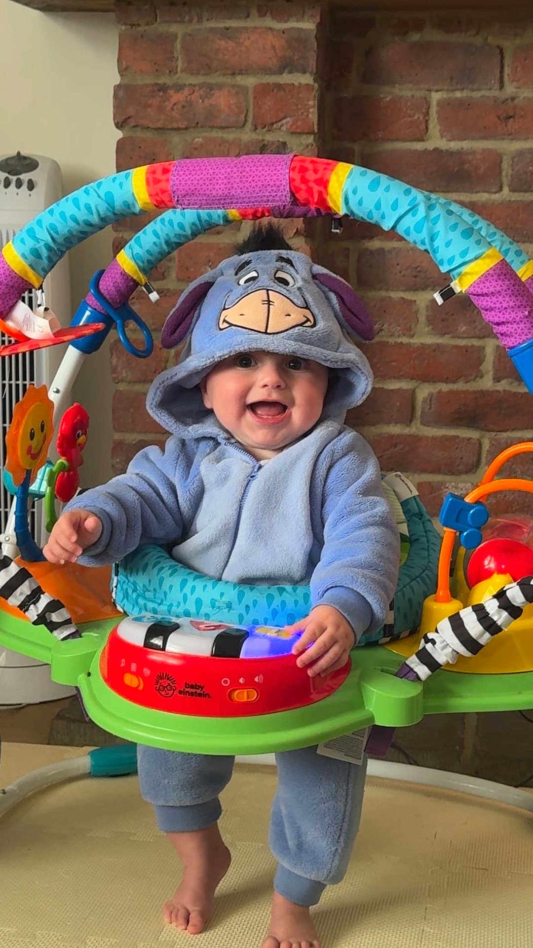 Ezra is registered to the contest to win money with this photo: baby, child, blue_onesie, eeyore_costume, baby_jumper, toy, colorful, brick_wall, happy, smiling, indoor, feet, playtime, cute, infant, hooded_clothing, person, early_childhood, home, entertainment
