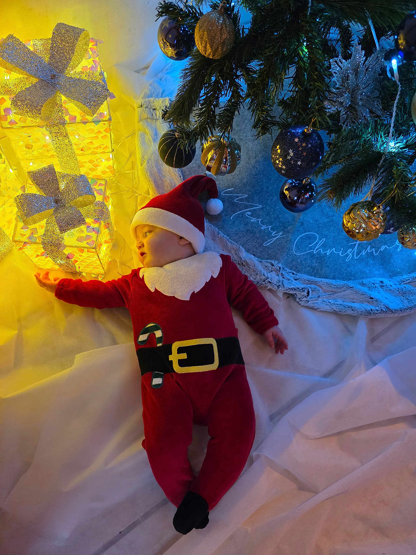 Matias a rejoint le concours — aidez-le/la à gagner de superbes lots ! baby, christmas, holiday, santa_claus_costume, gift_boxes, decorations, christmas_tree, ornaments, lights, festive, toddler, indoors, red_clothing, white_fabric, face, sleeping, floor, cozy, seasonal, celebration