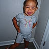 toddler, child, smile, posing, indoor, grey_outfit, shorts, tshirt, sandals, jewelry, necklace, braided_hair, happy, standing, floor, wall, corner, person, footwear, portrait