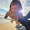 baby, child, jacket, hood, beach, sand, ocean, waves, sunlight, lens_flare, sky, clouds, person, hand, playing, outdoor, happy, smiling, warm_clothing, rocks