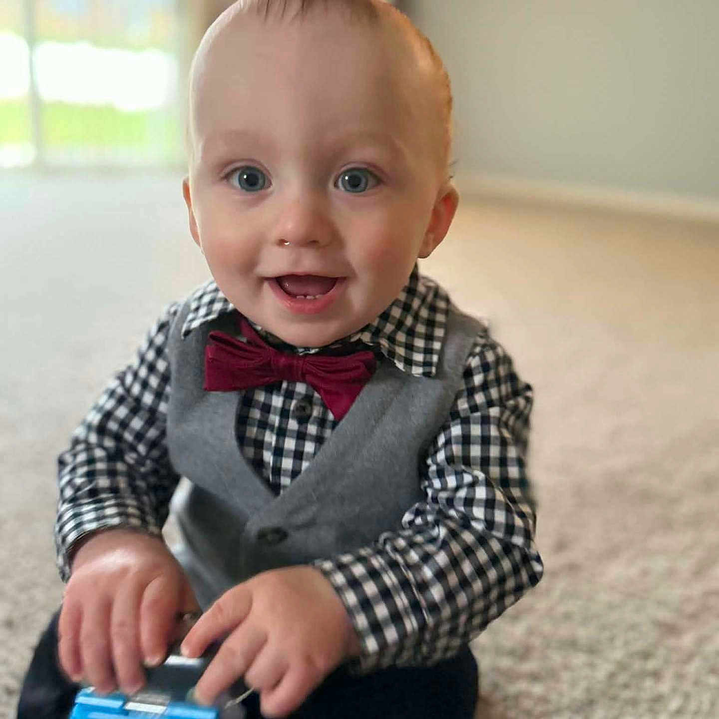 Jettson is registered to the contest to win money with this photo: toddler, child, boy, smiling, happy, indoor, carpet, toy, blue_toy, vest, bow_tie, checkered_shirt, hands, face, eyes, nose, mouth, hair, sitting, background_blur