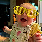 April joined the competition — help win amazing prizes! baby, child, infant, portrait, face, smiling, laughing, sunglasses, shutter_shades, yellow_glasses, indoor, home, floral_bib, yellow_clothing, outstretched_hand, reaching, closeup, funny, happy, photography