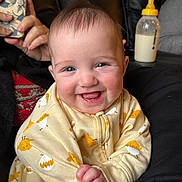 Chloe joined the competition — help win amazing prizes! baby, smiling, onesie, yellow, chick_print, infant, happy, face, hand, bottle, milk, person, phone, couch, black_clothing, indoor, portrait, cute, child, young