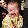 baby, smiling, onesie, yellow, chick_print, infant, happy, face, hand, bottle, milk, person, phone, couch, black_clothing, indoor, portrait, cute, child, young