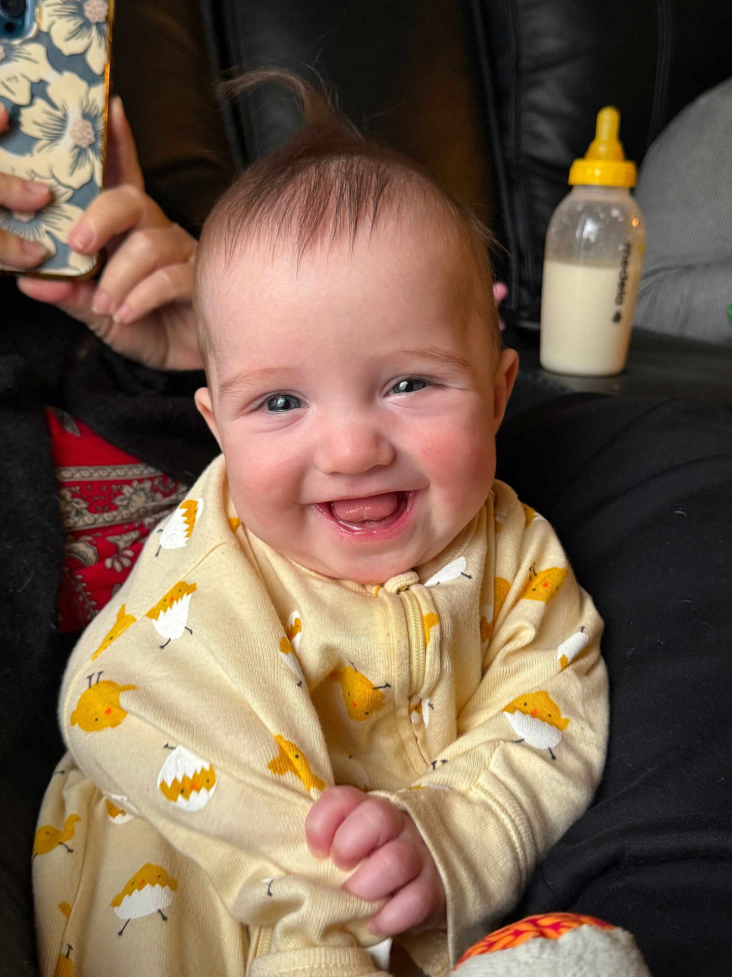 Chloe joined the competition — help win amazing prizes! baby, smiling, onesie, yellow, chick_print, infant, happy, face, hand, bottle, milk, person, phone, couch, black_clothing, indoor, portrait, cute, child, young
