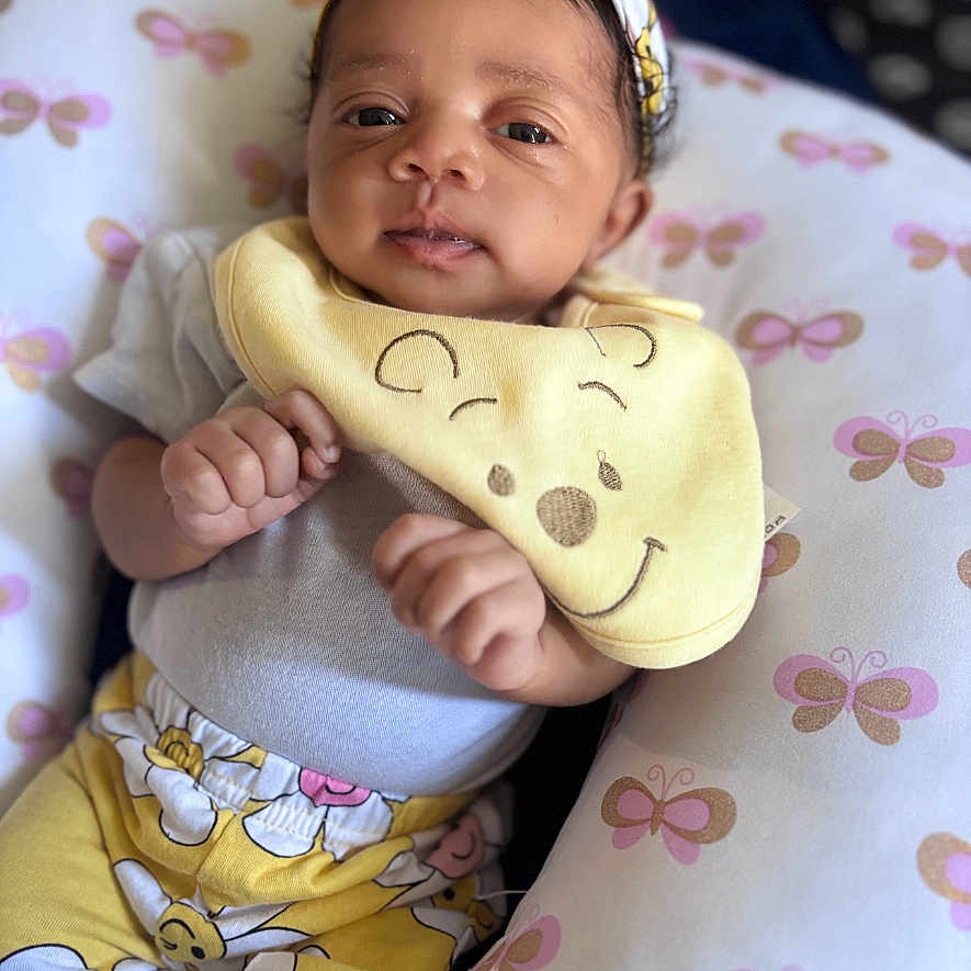 Nova is registered to the contest to win money with this photo: baby, bib, butterfly_pattern, child, clothing, comfort, cute, eyes, face, floral_pattern, hands, headband, indoor, infant, lying_down, pillow, portrait, smiling_face_bib, soft_toys, yellow_clothing