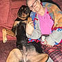 animal, blanket, brick_wall, carpet, casual_clothing, comfort, companion, couch, cozy, dog, friendship, glasses, home, indoor, person, pet, plaid_pants, relaxing, seated, smiling