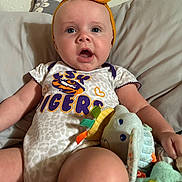 Addison is registered to the contest to win money with this photo: baby, child, onesie, headband, bow, plush_toy, elephant_toy, couch, pillow, indoor, person, infant, clothing, cute, expression, seated, soft_toy, toddler, wall, fabric