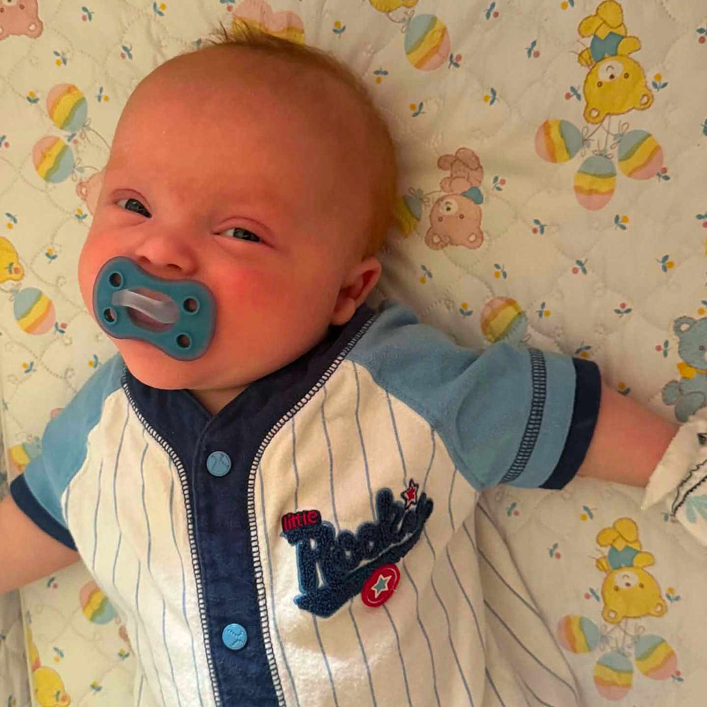 Braxton joined the competition — help win amazing prizes! baby, infant, child, pacifier, smiling, face, portrait, lying_down, blanket, crib_bedding, baby_clothing, baseball_outfit, striped_clothing, blue, colorful, teddy_bears, nursery, indoors, close_up, happy