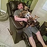 aquarium, beard, brown_dog, casual, companion, dog, furniture, home, indoor, living_room, man, multiple_dogs, pet_bed, recliner, relaxing, seated, shorts, small_dog, tile_floor, white_dog