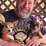 beard, brown_dog, casual_clothing, dog, friendship, glass_table, happy, holding, lattice, man, necklace, outdoor, pet, portrait, reflection, seated, small_dog, smiling, tan_dog, wooden_background