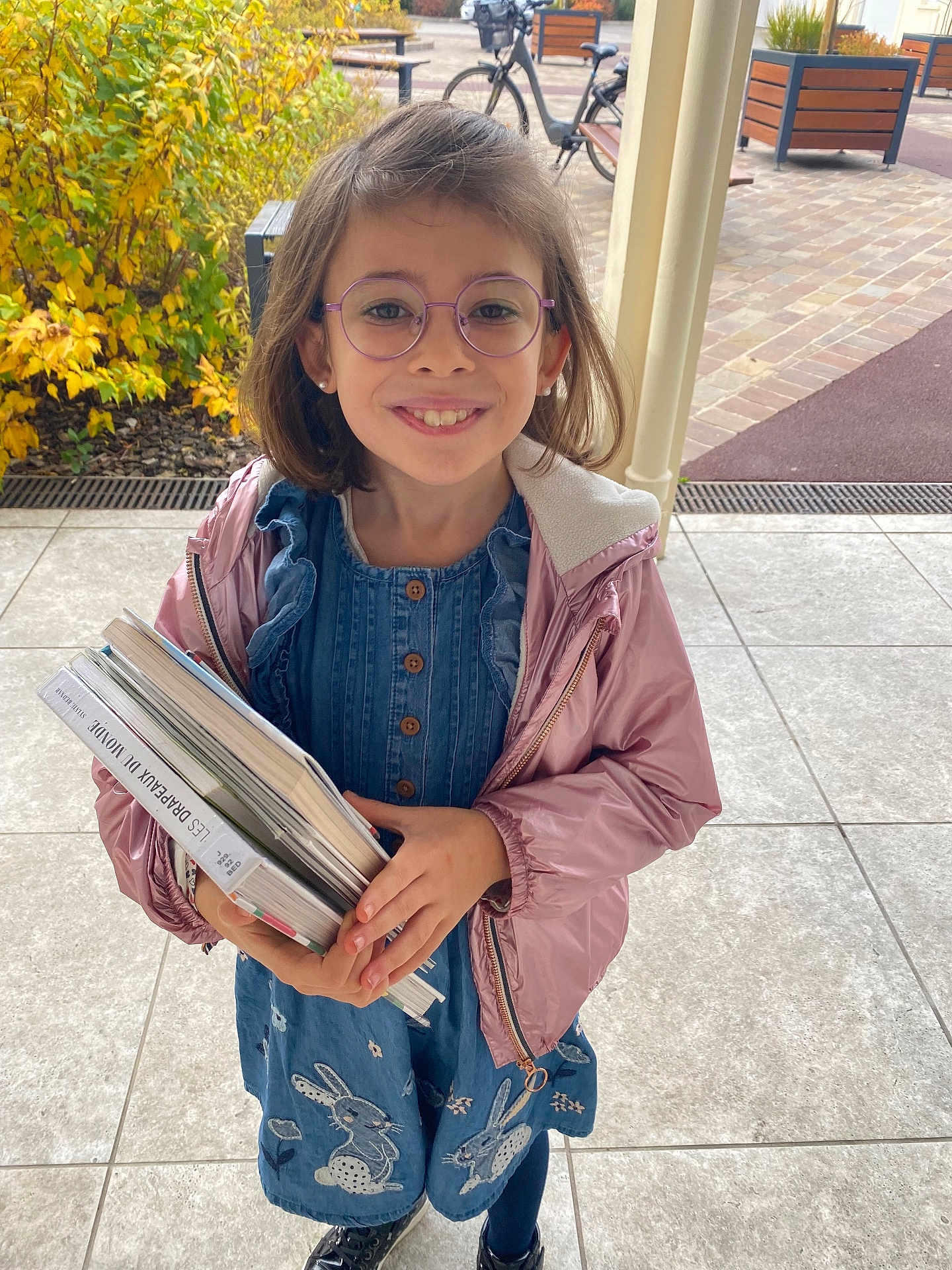 Chloé a rejoint le concours — aidez-le/la à gagner de superbes lots ! child, girl, glasses, smiling, books, blue_dress, pink_jacket, outdoor, autumn, bunnies, tiles, bicycle, plant, curly_hair, portrait, casual_clothing, happy, standing, daylight, young