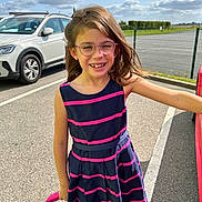 Chloé participe au concours pour gagner de l'argent avec cette photo : child, girl, glasses, smiling, dress, striped_clothing, outdoor, parking_lot, car, sunny, daylight, asphalt, fence, happy, person, hair, holding_object, casual, fashion, playful