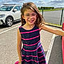 child, girl, glasses, smiling, dress, striped_clothing, outdoor, parking_lot, car, sunny, daylight, asphalt, fence, happy, person, hair, holding_object, casual, fashion, playful