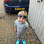 child, toddler, boy, smiling, sunglasses, blue_crocs, shorts, tshirt, car, volkswagen, gt, driveway, brick_pavement, shed, outdoor, grass, plants, feet, standing, daylight