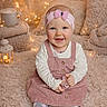 baby, child, girl, smiling, headband, pink_dress, long_sleeve_shirt, socks, indoor, soft_lighting, fairy_lights, blanket, pillow, lantern, cozy, cute, portrait, happy, floor_seating, warm_colors