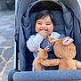 baby, stroller, smiling, plush_toy, toy, child, cuddly, happy, outdoor, pavement, cute, infant, clothing, seat, comfort, hand, face, person, head, fabric