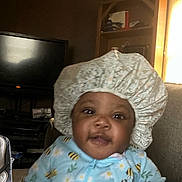 Princes Damiya is registered to the contest to win money with this photo: baby, child, bonnet, onesie, bee_pattern, indoor, living_room, television, shelf, furniture, curious, happy, person, smile, seated, cozy, home, infant, clothing, face