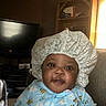 baby, child, bonnet, onesie, bee_pattern, indoor, living_room, television, shelf, furniture, curious, happy, person, smile, seated, cozy, home, infant, clothing, face