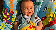 Stevie is registered to the contest to win money with this photo: baby, child, smiling, infant, seat, colorful, toy, fish, underwater_theme, comfort, happy, indoor, clothing, blanket, room, sunlight, face, person, sitting, playful