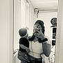 baby, child, woman, mirror, selfie, phone, headband, bow, black_and_white, indoor, room, curly_hair, clothing, purse, furniture, dartboard, shelf, casual, portrait, holding