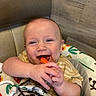 baby, child, high_chair, carrot, food, smile, happy, cute, infant, hands, face, chewing, clothing, pattern, fabric, indoor, wood, seat, playful, colorful