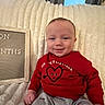 Kayson is registered to the contest to win money with this photo: baby, child, infant, smile, red_sweater, heart_design, gray_pants, blanket, couch, indoor, portrait, face, person, happy, seated, cozy, milestone_board, furniture, home, casual