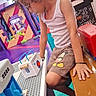 child, curly_hair, tank_top, shorts, mickey_mouse, toy, cash_register, play_area, bench, colorful, indoor, focused, hand, floor, wall_art, seat, game, kid, casual_clothing, pretend_play