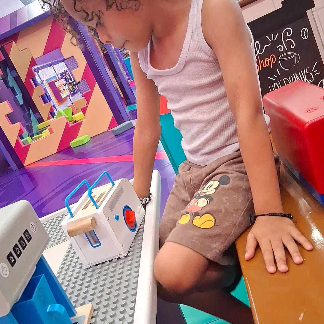 Da'Mani joined the competition — help win amazing prizes! child, curly_hair, tank_top, shorts, mickey_mouse, toy, cash_register, play_area, bench, colorful, indoor, focused, hand, floor, wall_art, seat, game, kid, casual_clothing, pretend_play