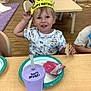 Meeko is registered to the contest to win money with this photo: toddler, birthday_crown, child, smiling, snack, cracker, purple_cup, table, chair, classroom, wooden_floor, plate, happy, person, indoor, clothing, short_sleeve_shirt, food, daycare, celebration
