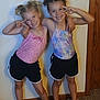 child, smiling, peace_sign, posing, shorts, sandals, indoor, wall, door_frame, carpet, anklet, toddler, happy, friends, hair_buns, colorful_clothing, footwear, two_people, standing, casual