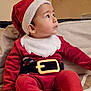 baby, child, santa_hat, red_clothing, belt, couch, indoor, curious, portrait, holiday_costume, young_child, seated, face, headwear, festive, cozy, looking_up, cute, expression, home