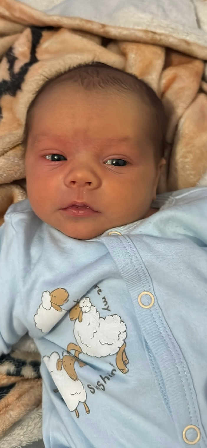 Lachlan is registered to the contest to win money with this photo: baby, infant, face, blanket, clothing, blue_outfit, sheep_print, cute, soft_texture, newborn, lying_down, portrait, child, warm_colors, cozy, peaceful, indoors, closeup, expression, sleepwear