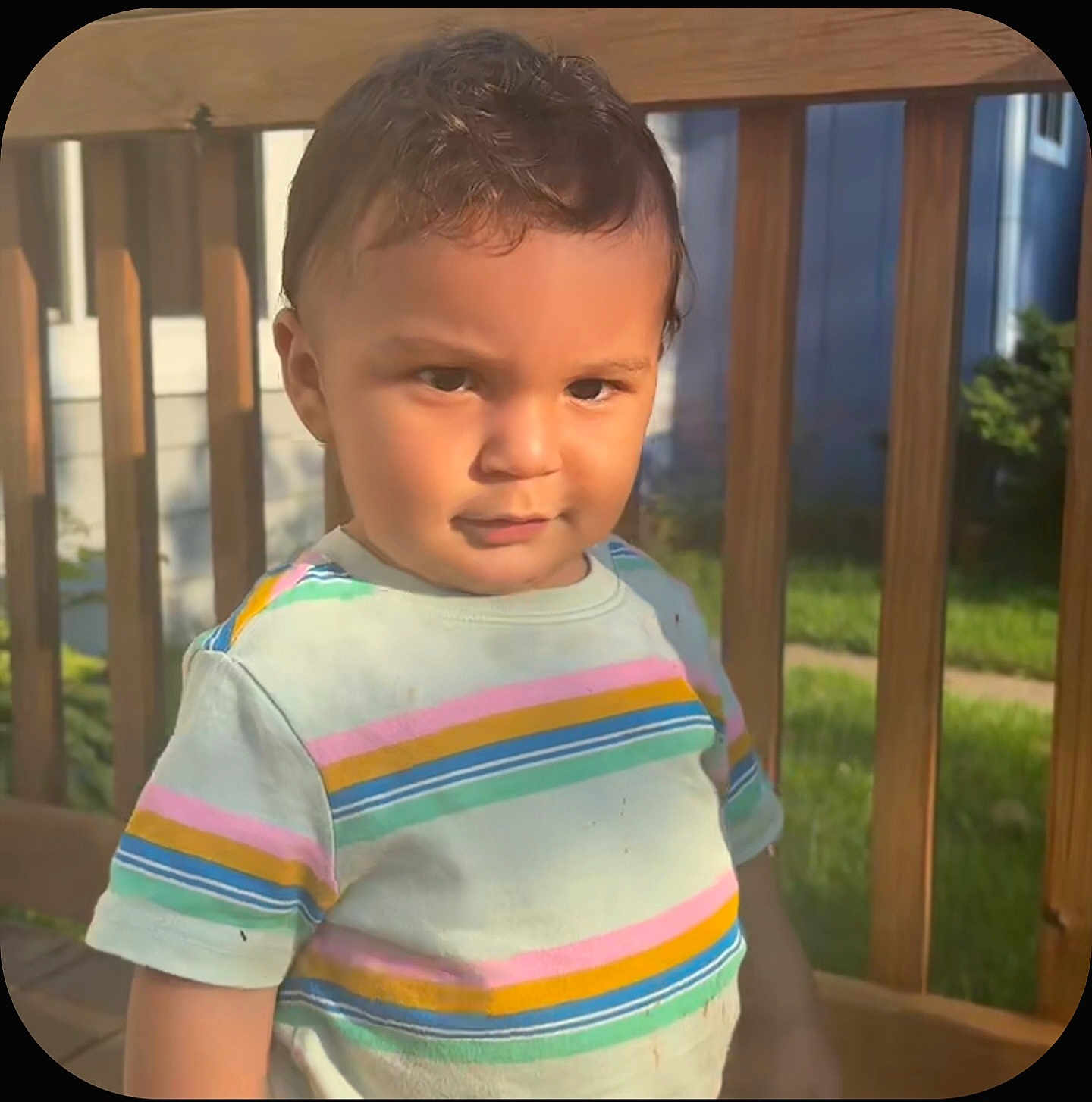 Koda is registered to the contest to win money with this photo: child, toddler, boy, outdoor, sunlight, striped_shirt, wooden_deck, railing, grass, portrait, smirk, short_hair, casual_clothing, daylight, person, nature, summer, expression, standing, curious