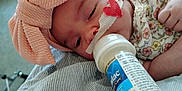 Rosie is registered to the contest to win money with this photo: baby, infant, bottle, feeding, headwrap, pink, bow, medical_tape, heart, floral_clothing, striped_cushion, close_up, face, hand, resting, indoors, cute, peaceful, portrait, child