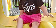 Colson is registered to the contest to win money with this photo: child, smiling, playground, slide, yellow_slide, black_tshirt, pink_shorts, sneakers, outdoor, daylight, fence, happy, casual_clothing, young_boy, play, fun, grass, metal_bars, person, feet