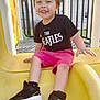 child, smiling, playground, slide, yellow_slide, black_tshirt, pink_shorts, sneakers, outdoor, daylight, fence, happy, casual_clothing, young_boy, play, fun, grass, metal_bars, person, feet