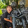 child, boy, statue, bench, outdoor, reading, book, sculpture, greenery, tree, happy, smiling, shorts, tshirt, footwear, nature, daylight, playful, public_art, park