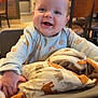 baby, child, high_chair, blanket, animal_print, smiling, happy, indoor, restaurant, wooden_chairs, table, face, person, clothing, cozy, cute, infant, joyful, portrait, seated