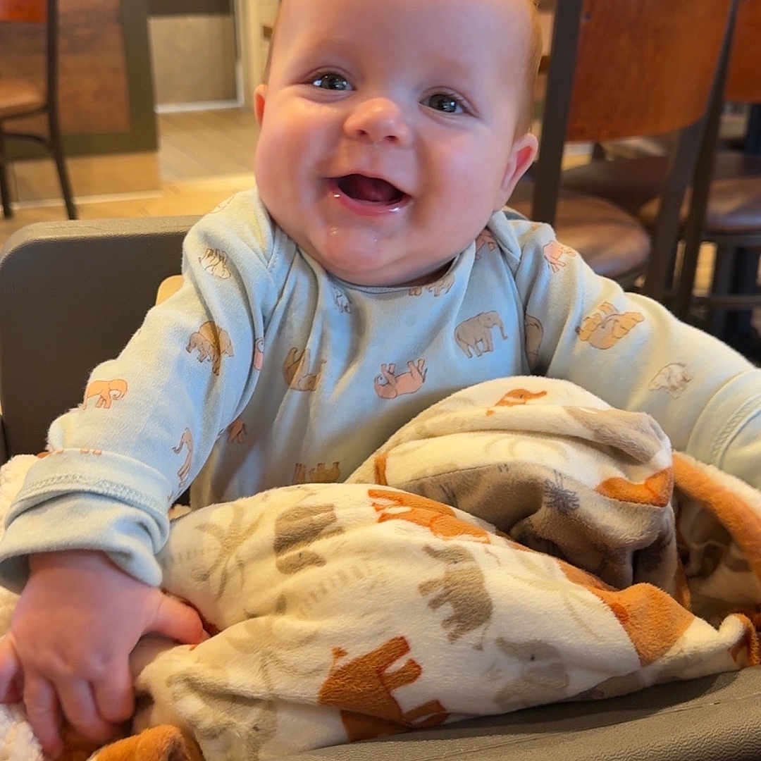 Aidyn is registered to the contest to win money with this photo: animal_print, baby, blanket, child, clothing, cozy, cute, face, happy, high_chair, indoor, infant, joyful, person, portrait, restaurant, seated, smiling, table, wooden_chairs