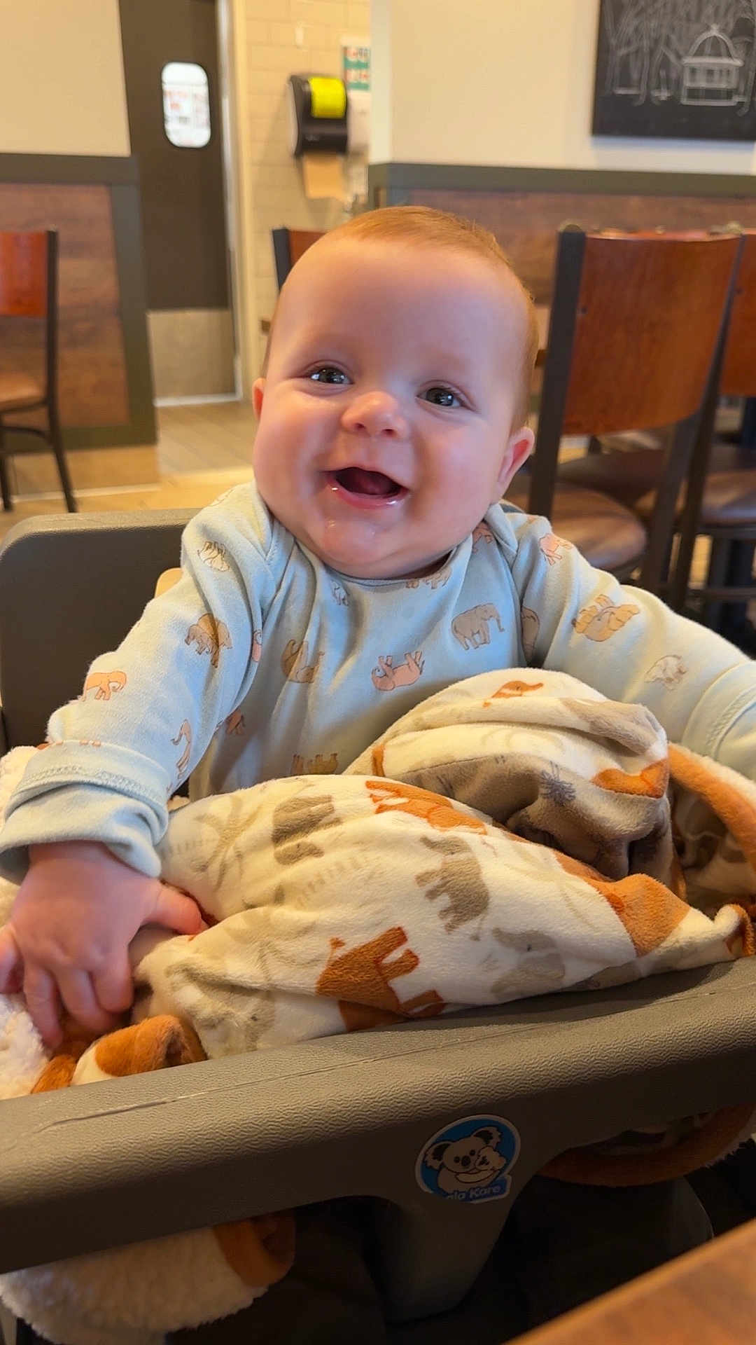 Aidyn is registered to the contest to win money with this photo: baby, child, high_chair, blanket, animal_print, smiling, happy, indoor, restaurant, wooden_chairs, table, face, person, clothing, cozy, cute, infant, joyful, portrait, seated