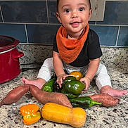Zion joined the competition — help win amazing prizes! baby, child, vegetables, sweet_potato, bell_pepper, butternut_squash, kitchen, countertop, orange_bib, black_shirt, white_pants, tile_wall, crock_pot, smiling, sitting, food, healthy, cute, indoors, curious