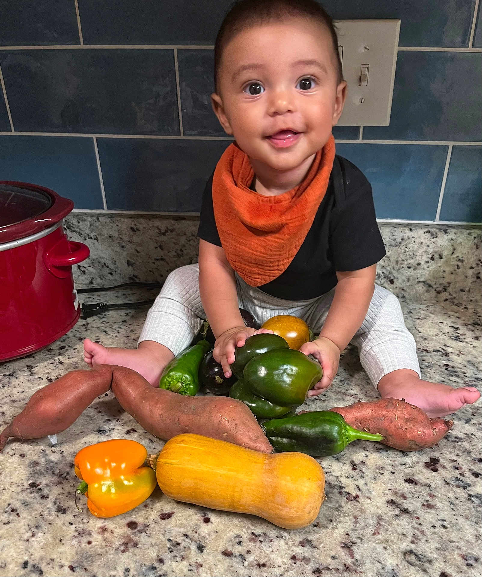 Zion joined the competition — help win amazing prizes! baby, child, vegetables, sweet_potato, bell_pepper, butternut_squash, kitchen, countertop, orange_bib, black_shirt, white_pants, tile_wall, crock_pot, smiling, sitting, food, healthy, cute, indoors, curious
