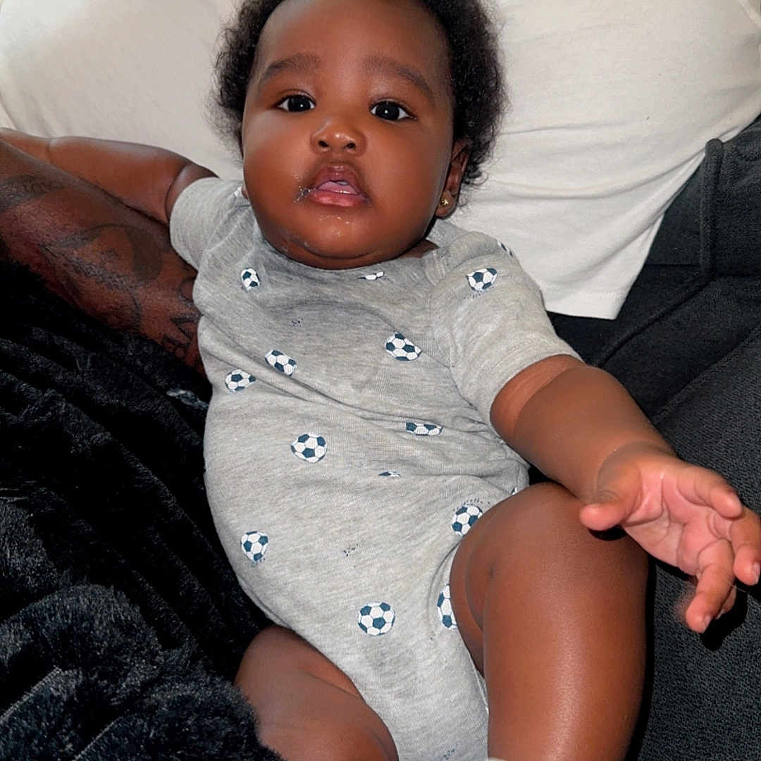 Trumias is registered to the contest to win money with this photo: baby, infant, child, man, person, couch, blanket, onesie, sock, hand, face, african_american, phone, tattoo, beard, sitting, reaching, indoor, relaxing, casual